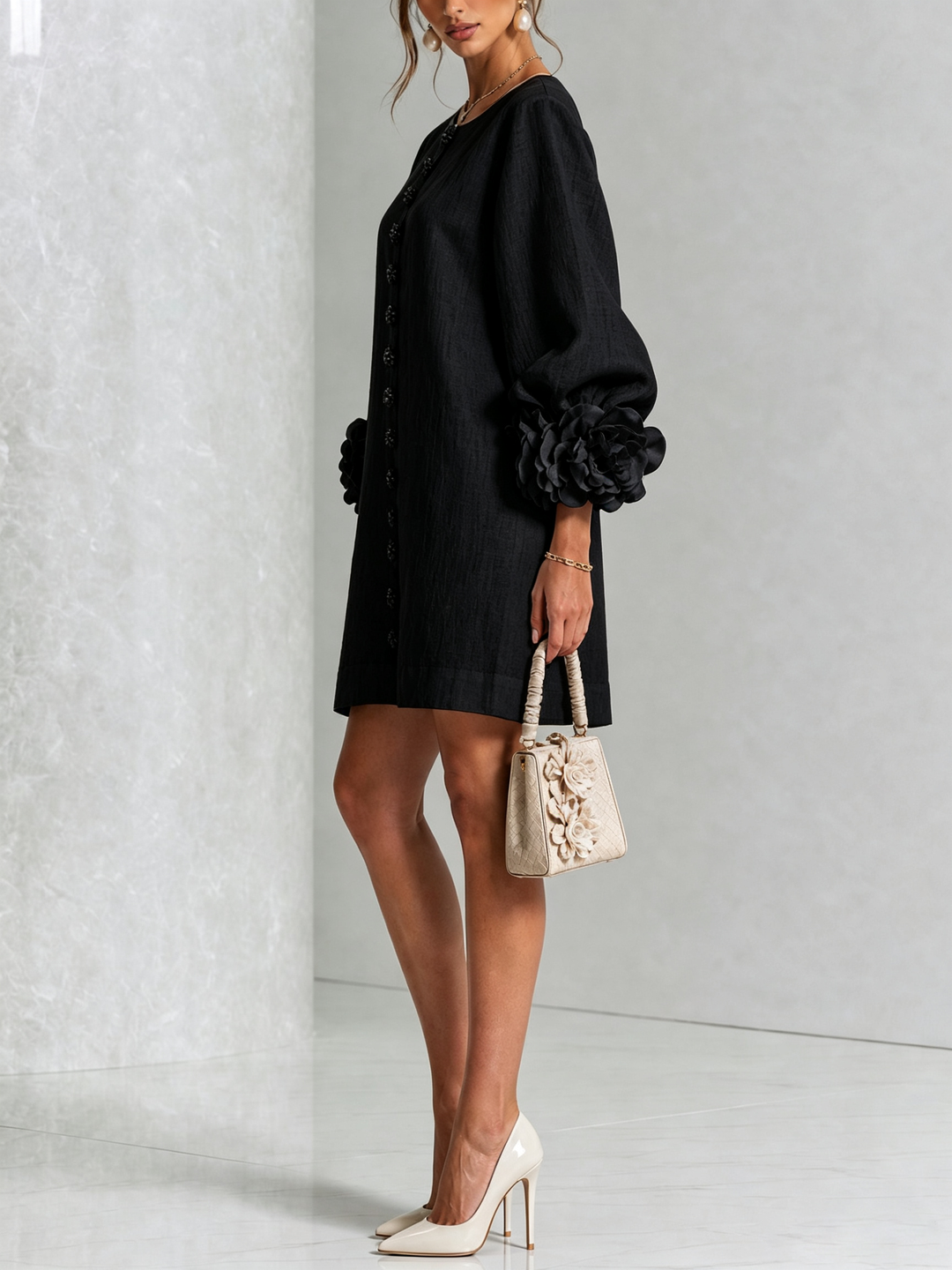 Robe noire portée de profil, manche ballon terminée par une grosse rose en tissu, avec sac à main beige et escarpins. 22
