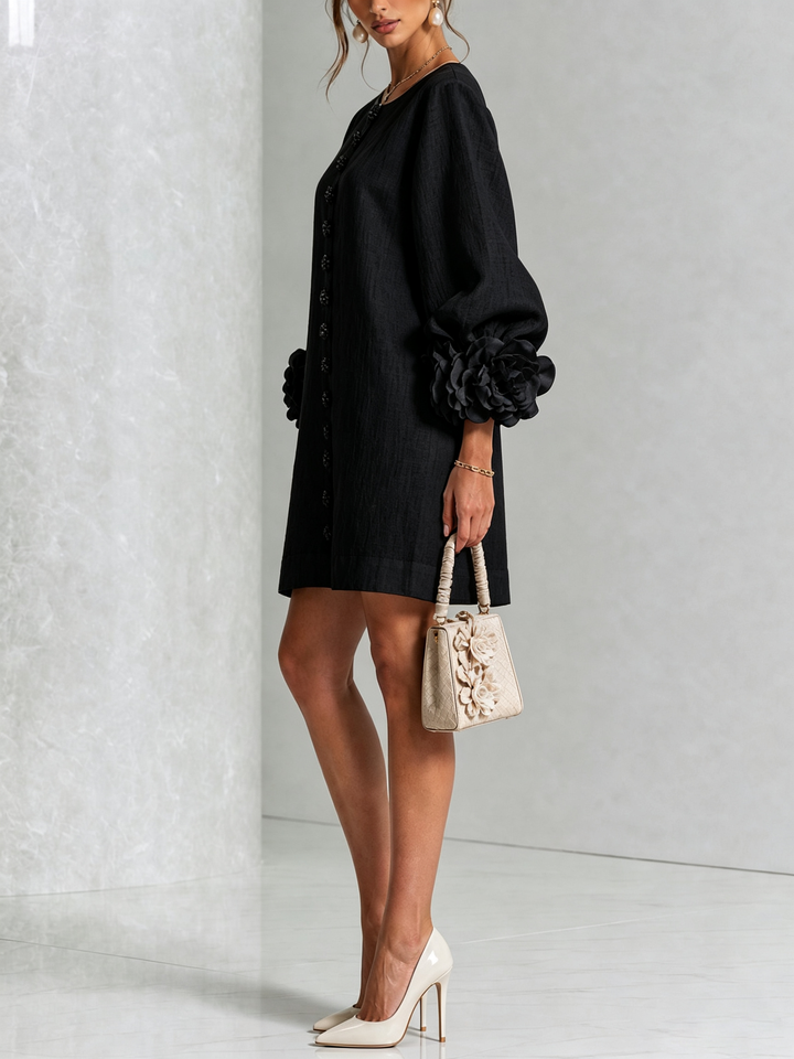 Robe noire portée de profil, manche ballon terminée par une grosse rose en tissu, avec sac à main beige et escarpins. 22