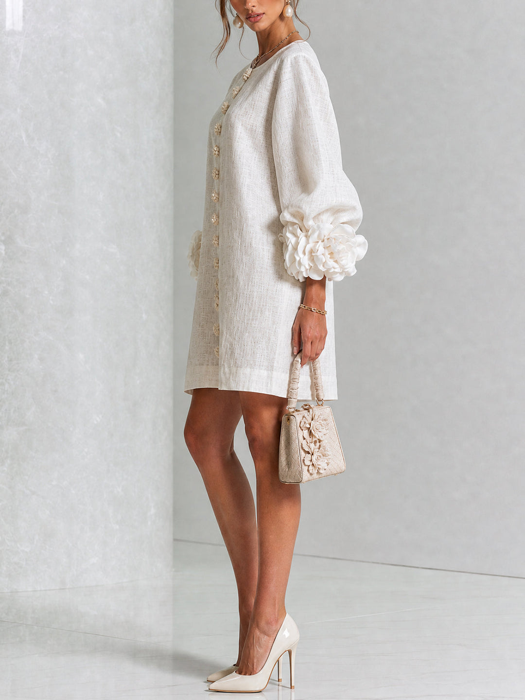 Robe crème portée de profil, manches bouffantes terminées par de grosses fleurs, avec sac à main beige et escarpins clairs. 2
