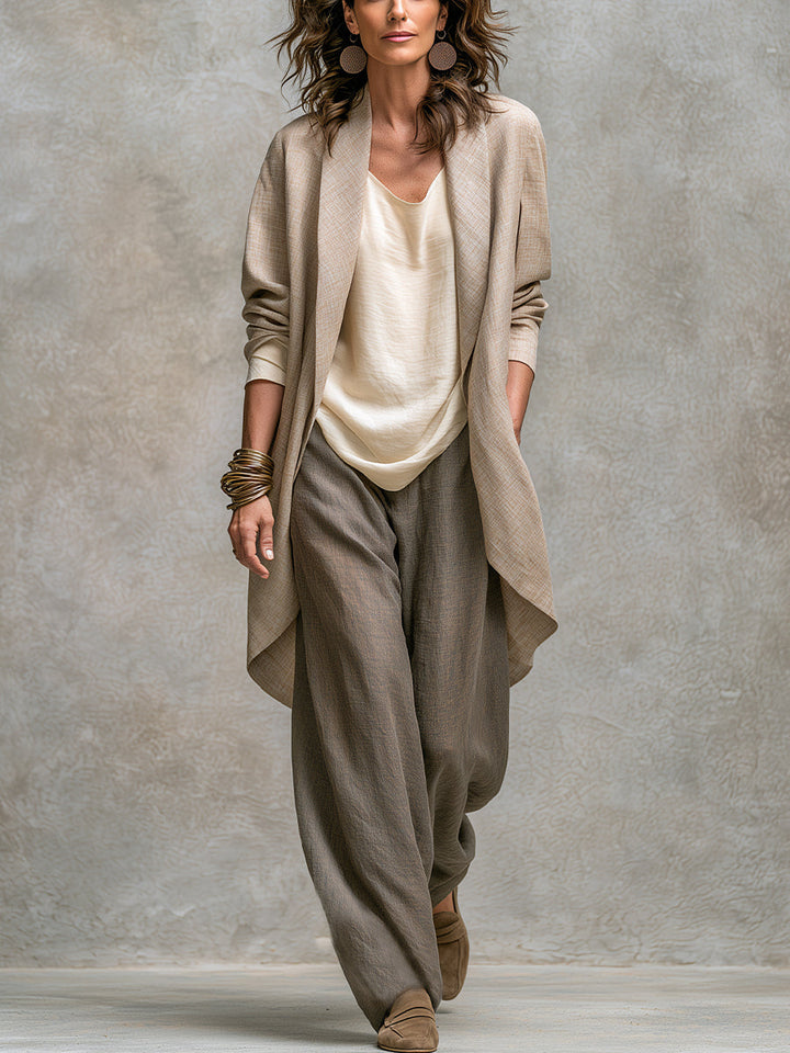 Femme en pied de face portant la longue veste ouverte beige sur le haut crème et le pantalon large taupe, devant un mur gris. 1