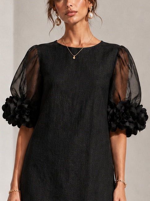Gros plan sur le haut de la robe noire texturée : encolure ronde, manches en voile transparent et fleurs en relief aux poignets. 5