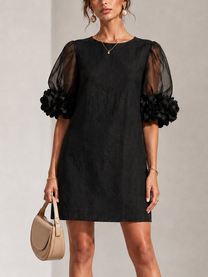 Robe noire droite portée de face, manches bouffantes transparentes terminées par de grosses fleurs en relief, avec un sac beige à la main. 1