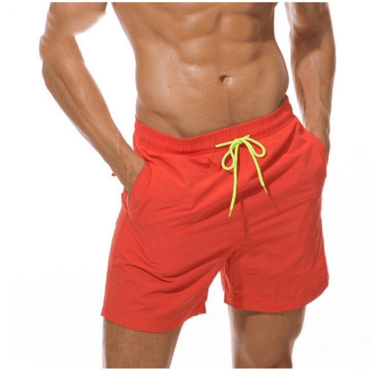 Short de bain rouge orangé porté, cordon jaune fluo, les deux mains dans les poches latérales. 6