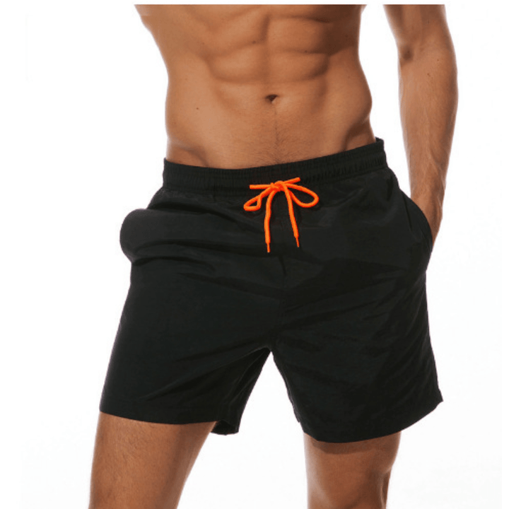 Short de bain noir porté par un mannequin, vue de trois quarts, cordon orange noué et une main dans la poche. 1