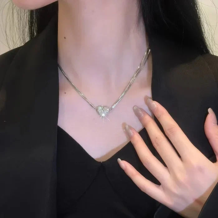 Collier à plusieurs fines chaînes argentées et cœur en strass porté au cou, avec une veste noire et une main posée sur la poitrine. 2