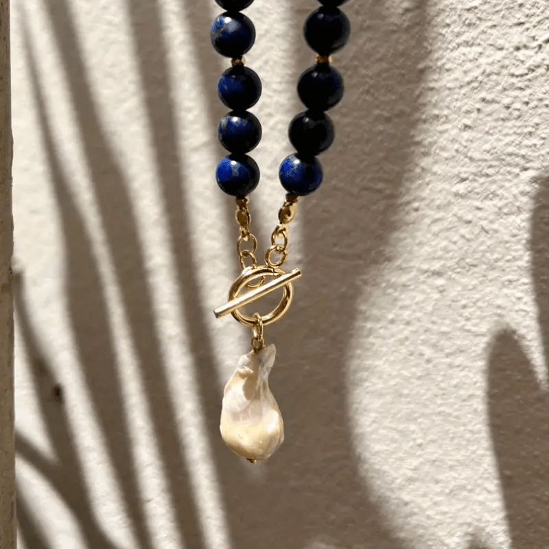 Pendentif en perle baroque et fermoir doré suspendus devant un mur blanc rayé d'ombres 8