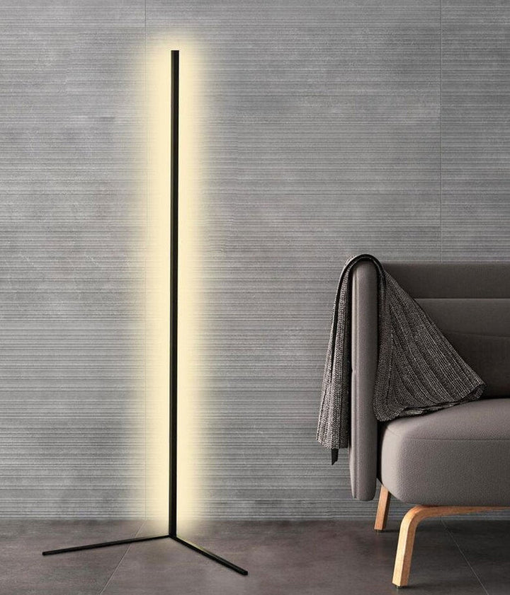 Lampadaire noir à pied en V, lumière blanc chaud, devant un mur gris strié et un fauteuil avec plaid 8