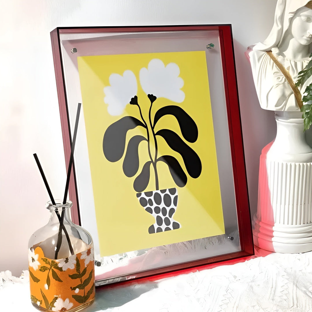 Cadre acrylique à bords rouge sombre à côté d'un diffuseur de parfum et d'un buste blanc, avec une illustration de plante noire sur fond jaune. 15