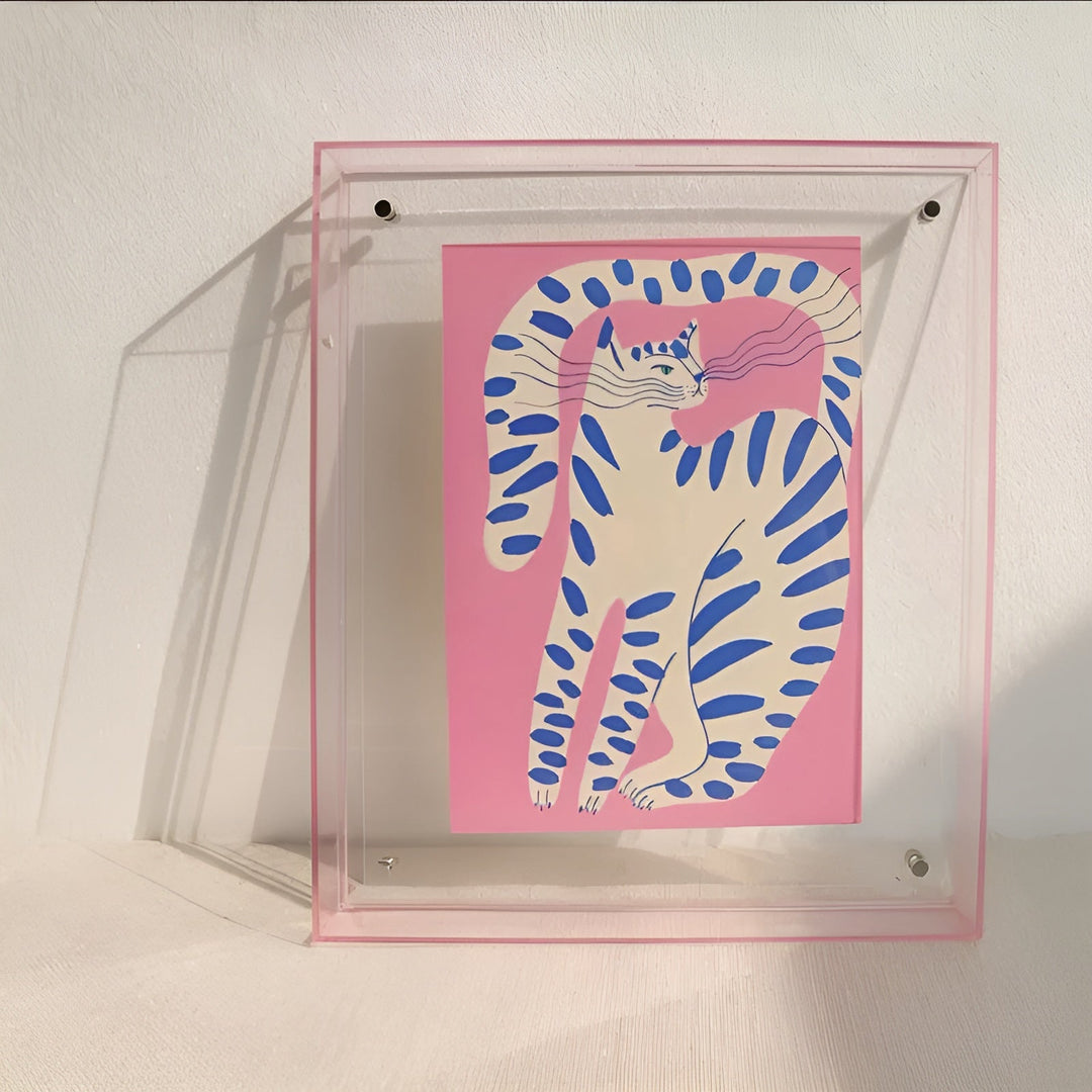 Cadre acrylique transparent à fine bordure rose appuyé contre un mur, avec une illustration de chat blanc rayé de bleu sur fond rose. 9