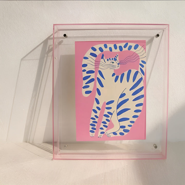 Cadre acrylique transparent à fine bordure rose appuyé contre un mur, avec une illustration de chat blanc rayé de bleu sur fond rose. 9