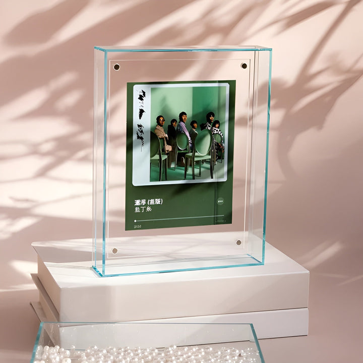 Cadre acrylique transparent à reflets turquoise posé sur des livres blancs, avec une pochette d'album vert, devant des ombres de feuillage. 13