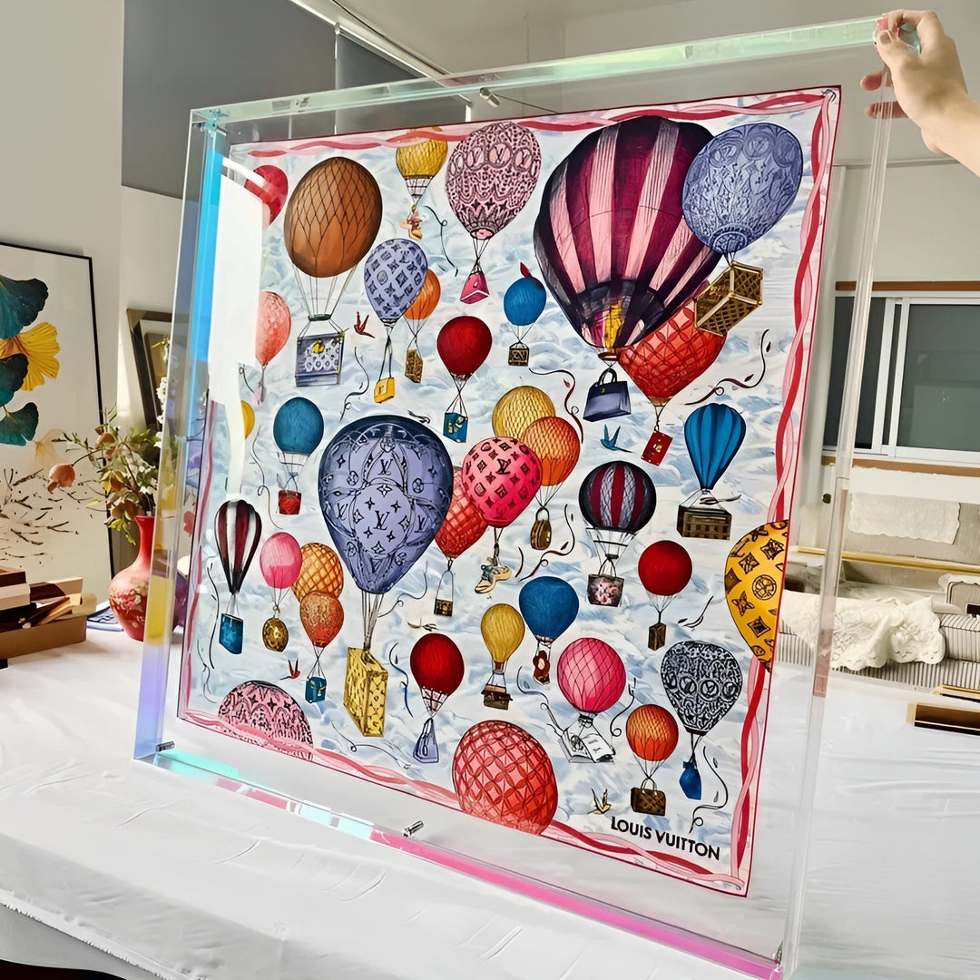 Très grand cadre acrylique transparent aux reflets irisés tenu à la main, contenant un foulard imprimé de montgolfières colorées. 11