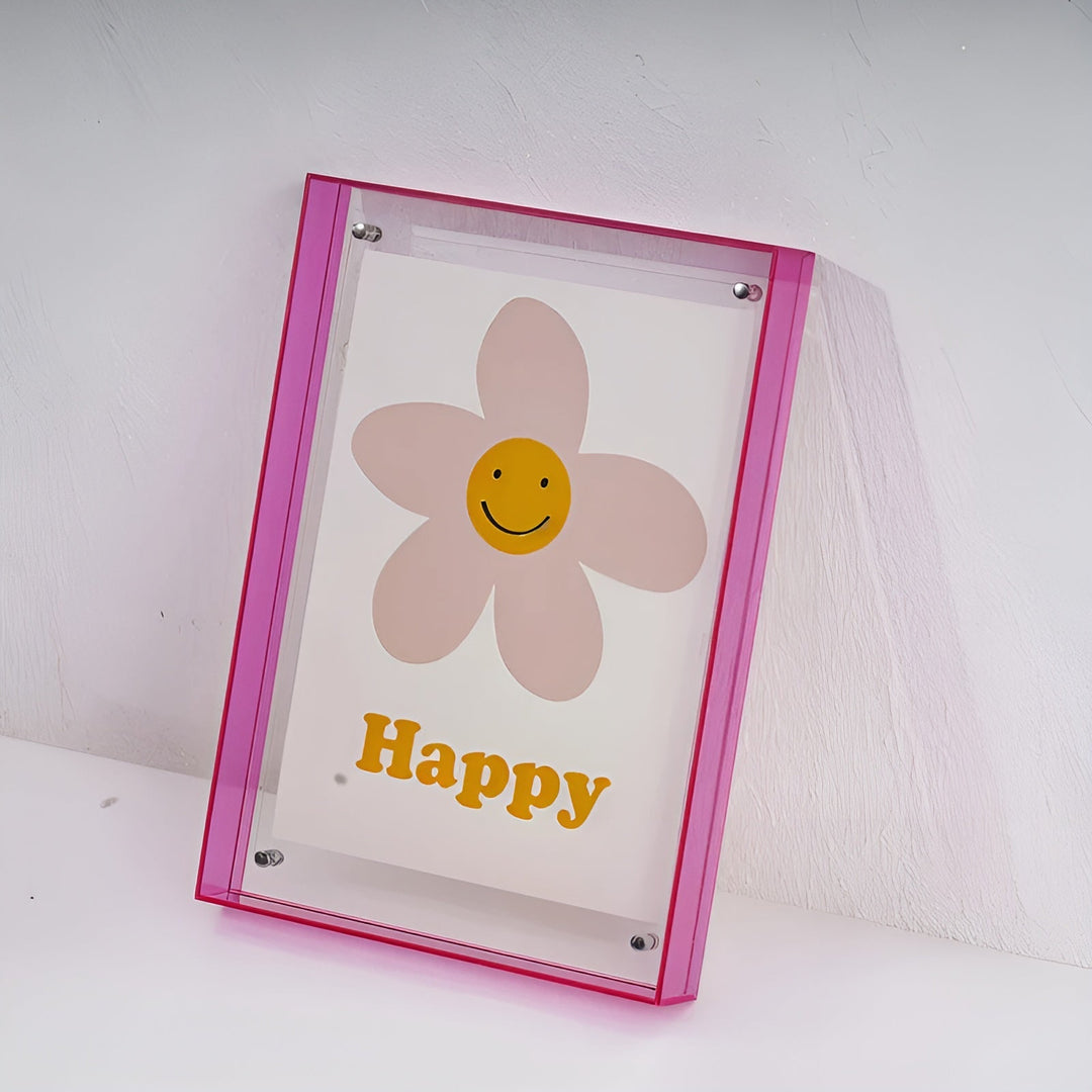 Cadre acrylique à bords rose vif appuyé contre un mur blanc, vu de trois quarts, avec l'illustration de fleur souriante. 19
