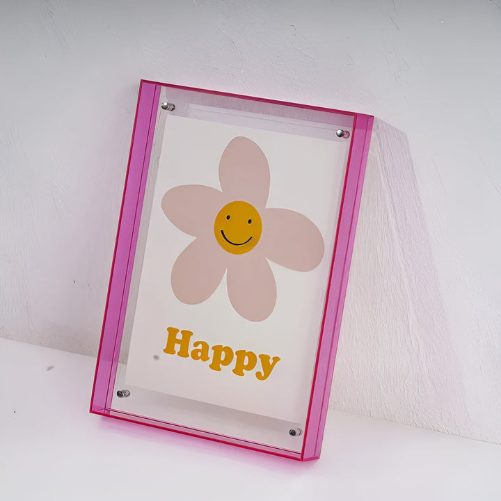 Cadre acrylique à bords rose vif appuyé contre un mur blanc, vu de trois quarts, avec l'illustration de fleur souriante. 19