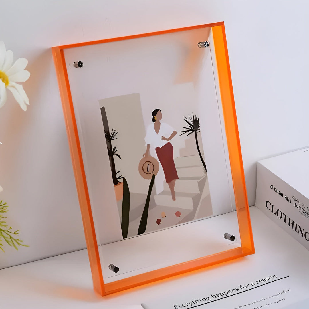 Cadre acrylique à bords orange vif sur une étagère, avec une illustration de femme à chapeau près de palmiers, à côté d'une fleur blanche. 10