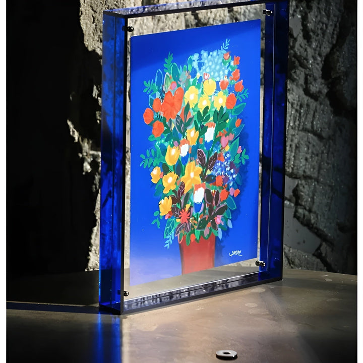 Cadre acrylique bleu intense posé sur une table devant un mur de pierre, avec un grand bouquet coloré sur fond bleu. 4
