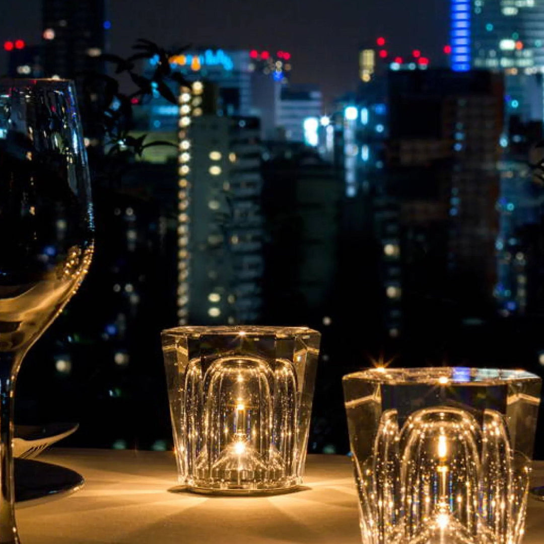 Deux lampes en cristal allumées sur une table au crépuscule, à côté d'un verre à pied, devant une vue nocturne de ville. 3