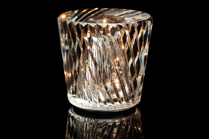 Lampe en cristal à facettes torsadées allumée sur fond noir, avec son socle foncé et son reflet en dessous. 12