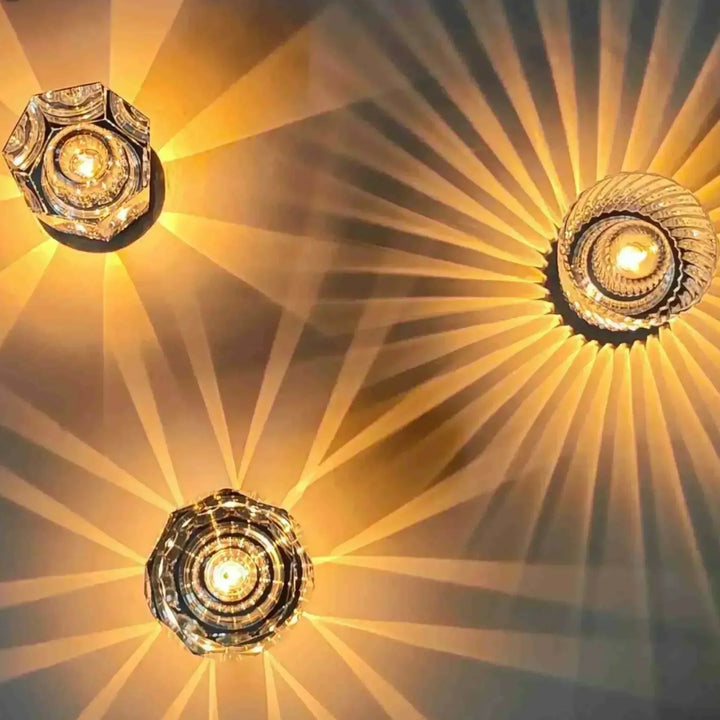 Vue de dessus de trois lampes en cristal allumées, aux formes hexagonale, ronde et à facettes, diffusant des rayons dorés. 2