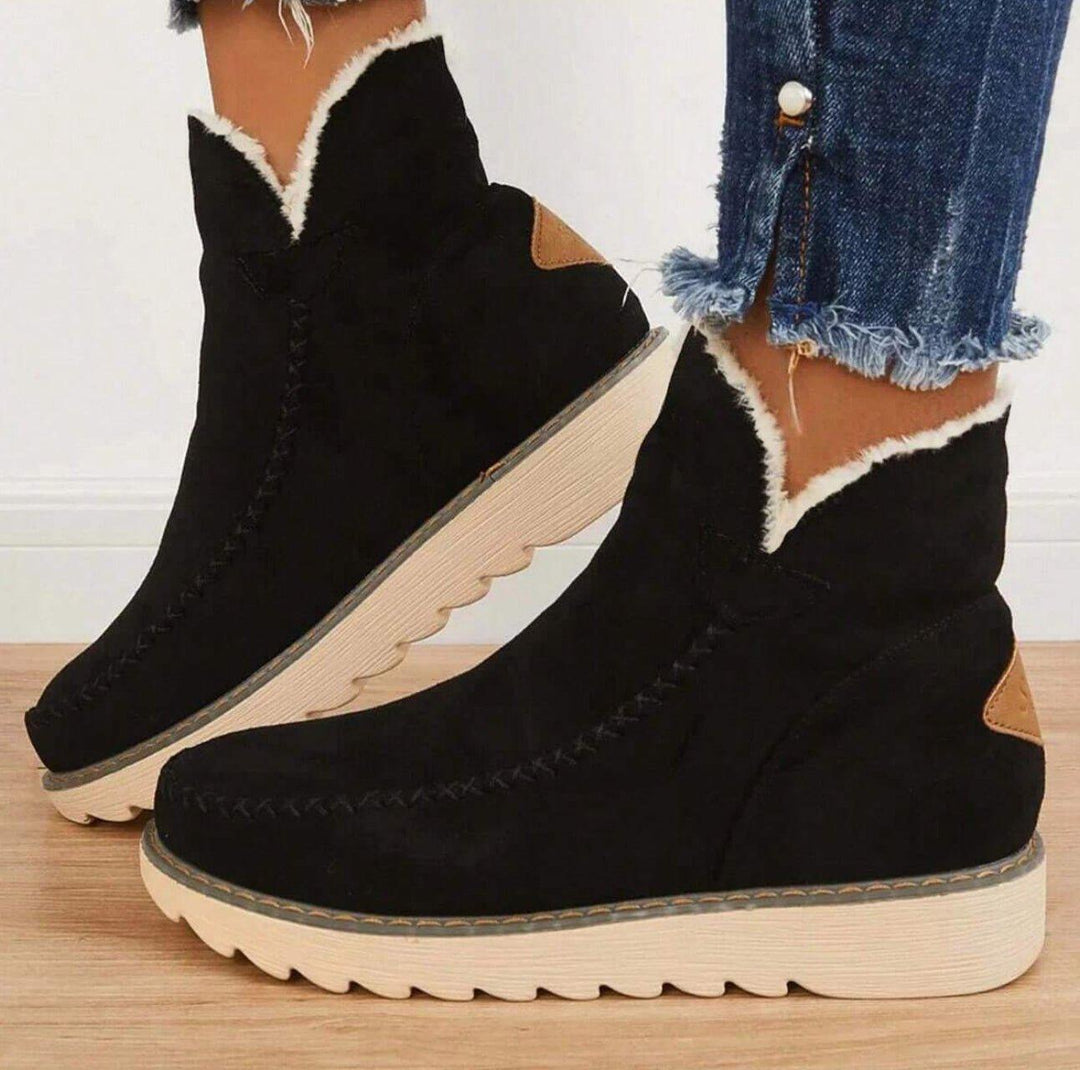 Vue de profil de bottines noires en suédine doublées de fourrure claire, portées avec un jean effiloché sur un parquet clair. 1