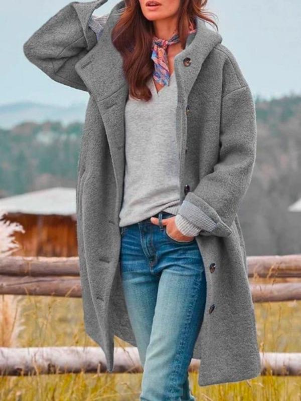 Manteau long à capuche gris porté ouvert sur un pull gris et un jean, foulard au cou, main relevée vers la tête. 2