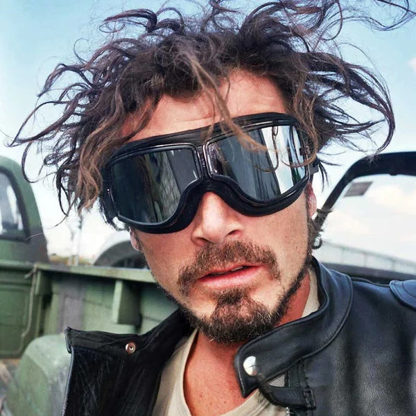 Homme aux cheveux ébouriffés portant les lunettes noires à verres miroir devant un véhicule vert, en blouson de cuir. 3