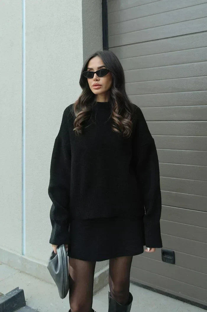 Ensemble noir porté en extérieur devant un mur beige et un portail gris, avec pochette grise et bottes noires. 5
