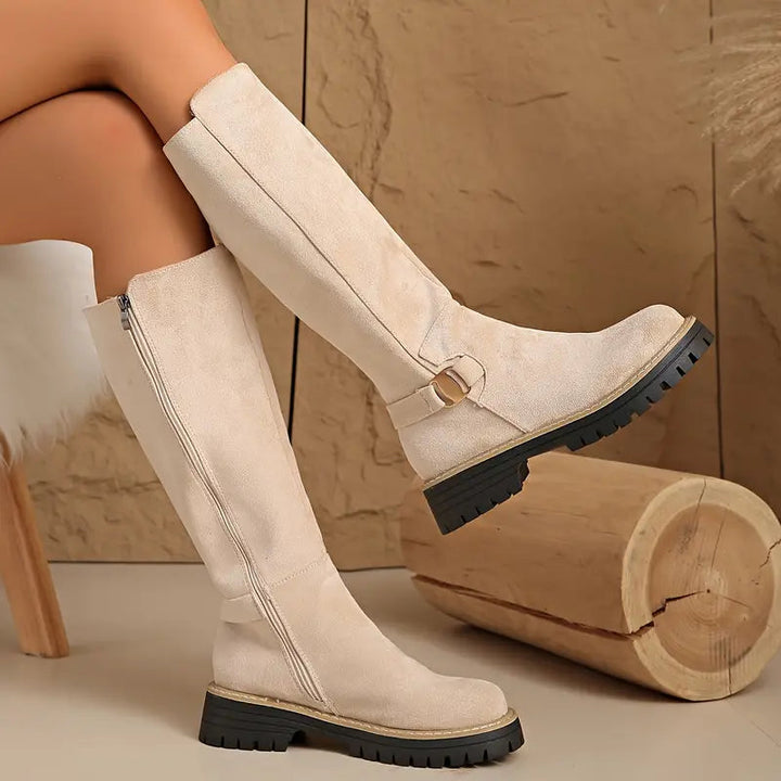 Bottes hautes beige clair en effet daim portées jambes croisées, avec fermeture éclair latérale et semelle noire épaisse. 8