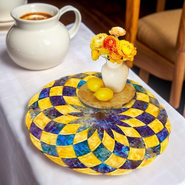 Set de table rond en patchwork jaune, violet et bleu sur une nappe blanche, avec un petit vase de fleurs et des citrons. 2