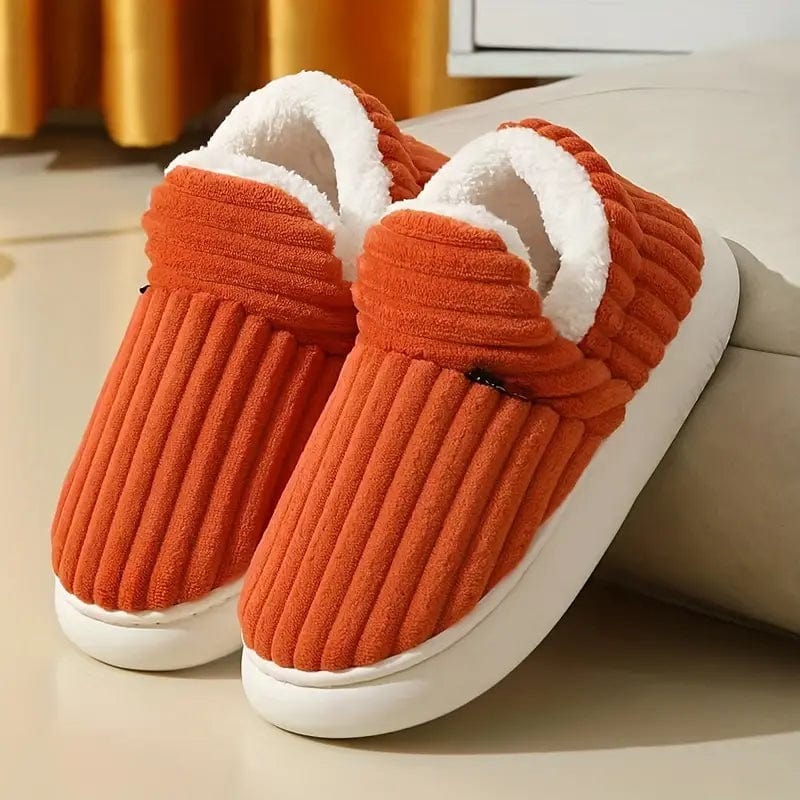 Paire de chaussons orange côtelés à doublure blanche douillette et semelle blanche, appuyée contre un canapé dans un intérieur chaleureux. 22