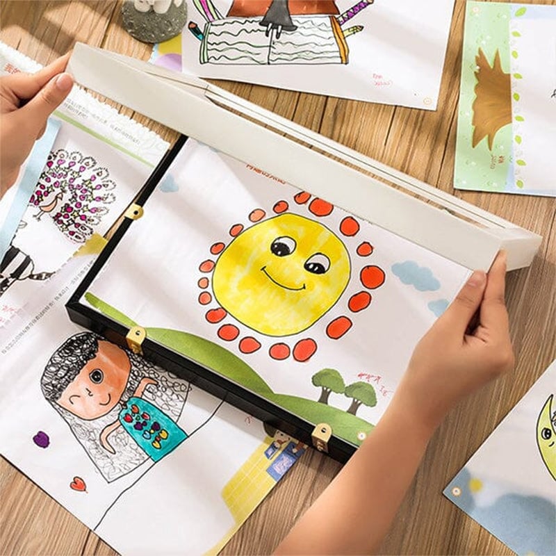 Mains d'enfant ouvrant un cadre noir sur une table en bois, au milieu de dessins colorés dont un soleil souriant. 12