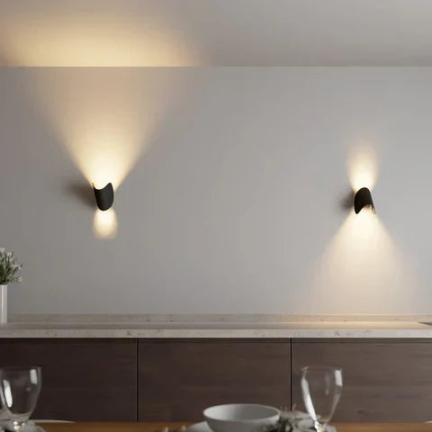 Deux appliques noires inclinées sur un mur clair au-dessus d'un meuble de salle à manger, éclairant en faisceaux vers le haut et le bas. 3