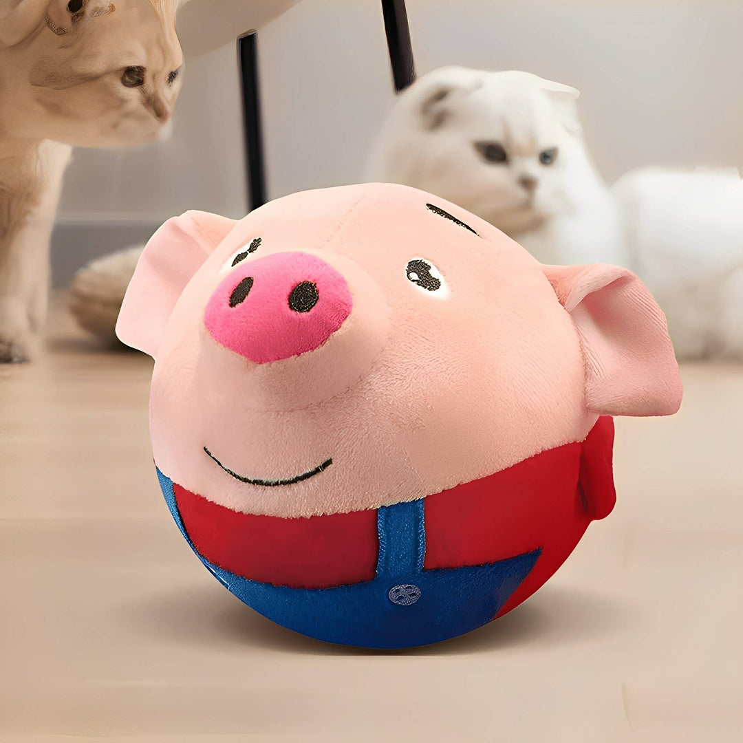 Balle en peluche cochon rose à salopette bleue et haut rouge, posée au sol, deux chats flous en arrière-plan. 3