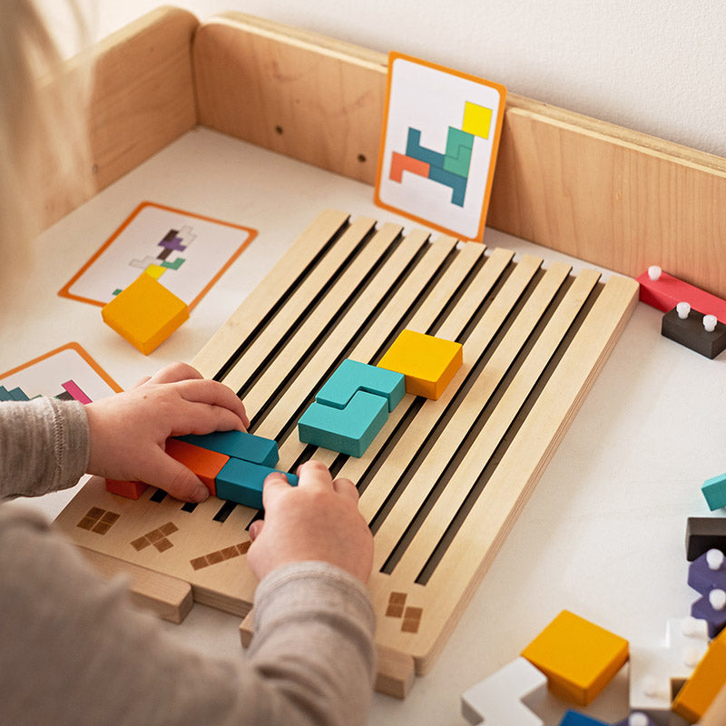 Mains d'un enfant glissant des pièces colorées dans les rainures du plateau posé sur une table, avec des cartes modèles derrière. 3