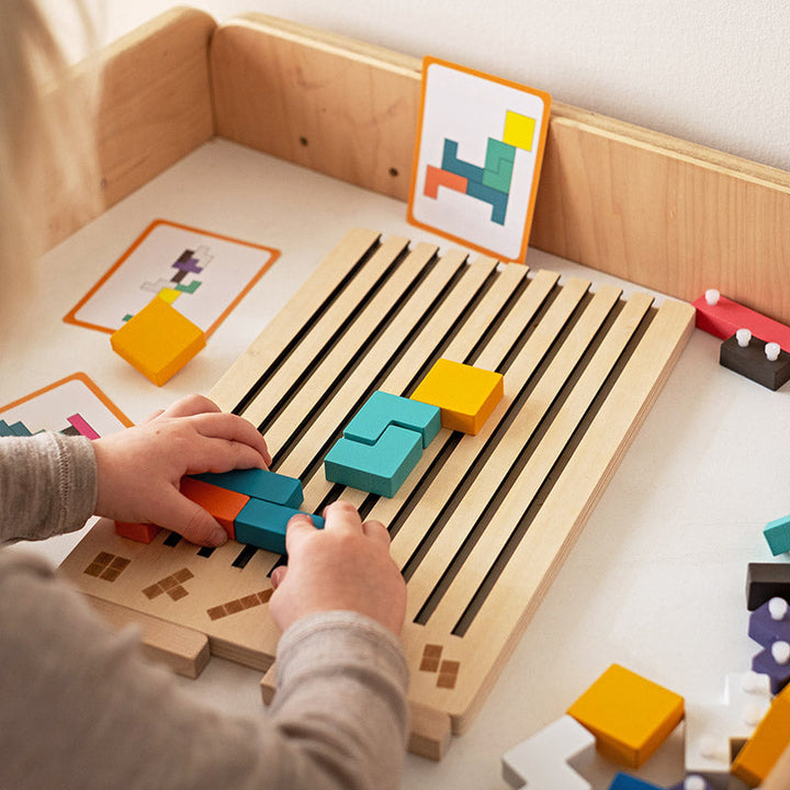 Mains d'un enfant glissant des pièces colorées dans les rainures du plateau posé sur une table, avec des cartes modèles derrière. 3