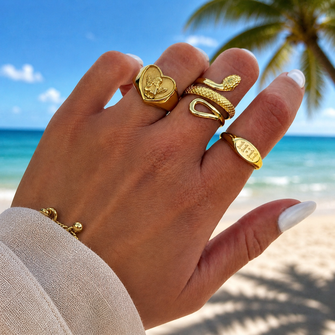 Main portant la bague cœur dorée avec ange, une bague serpent et une bague ovale gravée, devant une plage et un palmier. 3