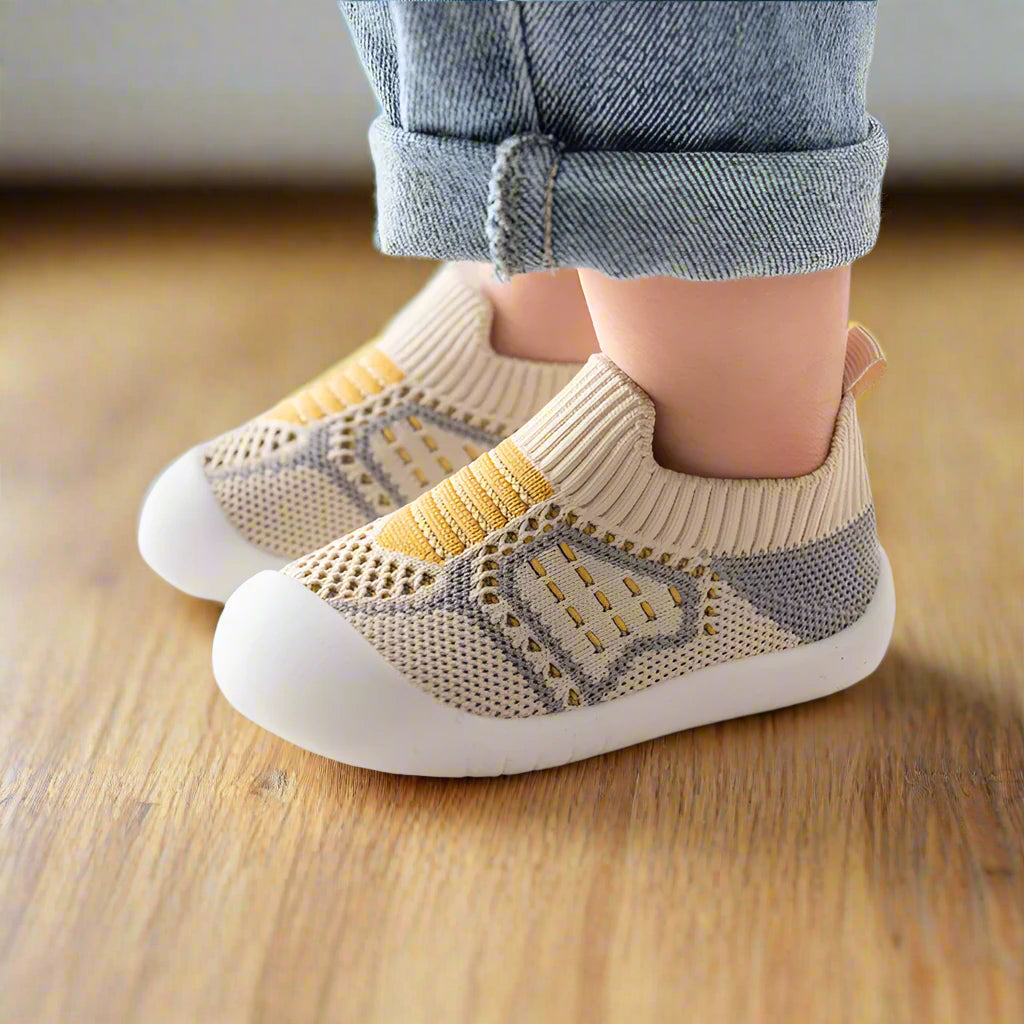 Vue de côté de chaussures en maille beige et grise à rayures jaunes, portées par un enfant sur un parquet en bois. 9