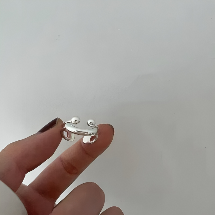 Bague argentée tenue entre deux doigts devant un mur gris clair, montrant ses deux boules et son arc lisse. 4