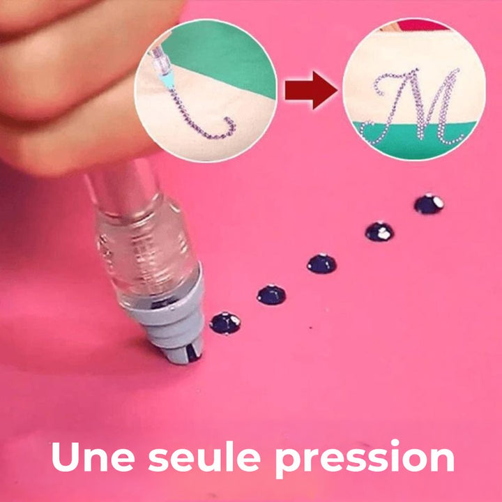 Stylo appliquant des strass sombres un à un sur un fond rose, avec lettre ornée en médaillon 3