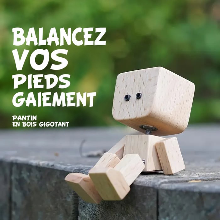 Personnage en bois aux jambes pendantes assis sur un rebord en pierre, devant un fond de verdure flou. 13
