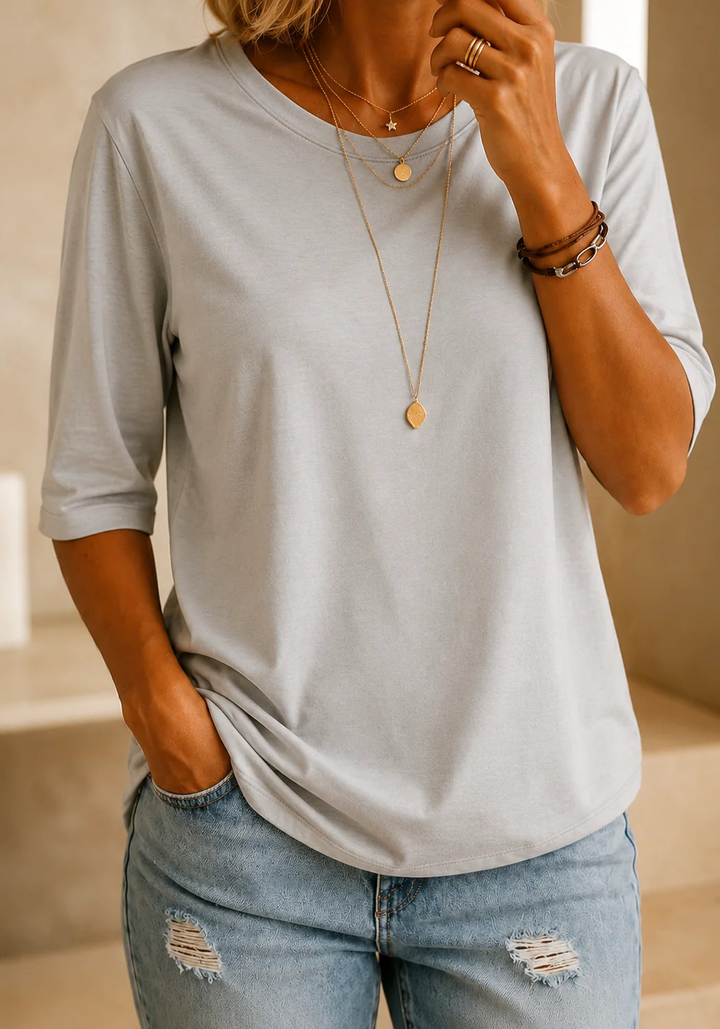 T-shirt gris clair à demi-manches porté avec un jean délavé et colliers dorés, dans un intérieur lumineux aux tons sable. 13