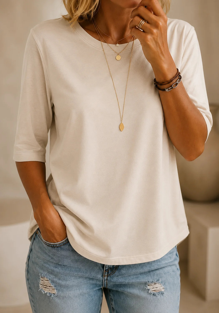 T-shirt crème à col rond et demi-manches porté avec un jean clair troué, devant un décor beige avec un vase. 12