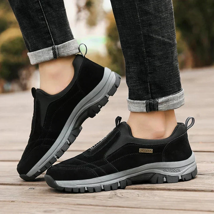 Chaussures noires portées par un homme en jean retroussé, debout sur un sol en bois, l'un des pieds légèrement levé. 4