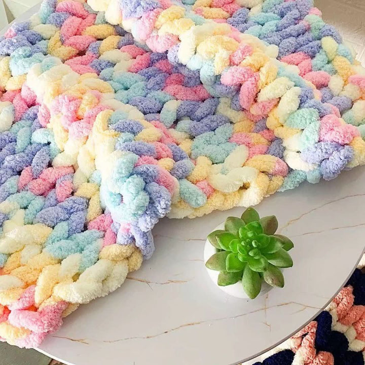Couverture en grosse maille multicolore aux tons pastel rose, bleu, jaune et violet, posée sur une table ronde en marbre avec une plante grasse. 1