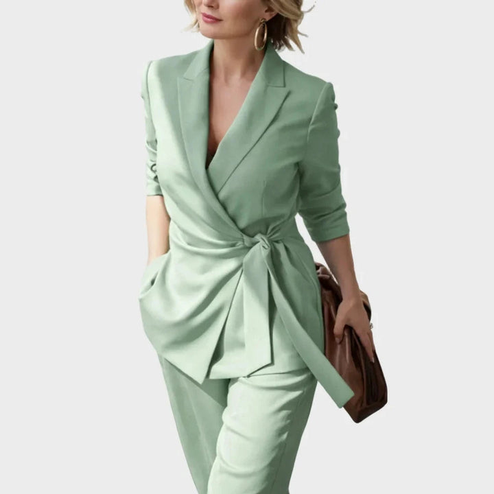 Gros plan en mouvement sur le blazer vert clair croisé et son nœud à la taille, porté par une femme blonde avec un sac marron. 6
