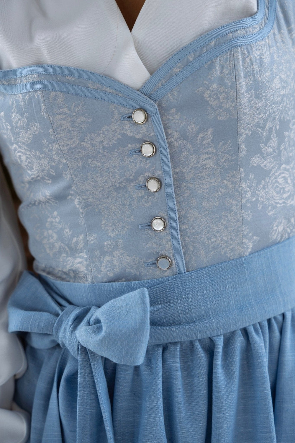Gros plan sur le corsage bleu à motif floral tissé, ses cinq boutons nacrés, la bordure en ruban et le nœud du tablier. 2