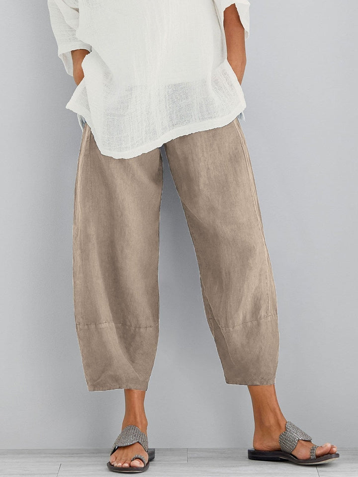 Pantalon large beige à taille élastique et bas resserré, porté avec une blouse blanche ample et des sandales sur fond gris. 2