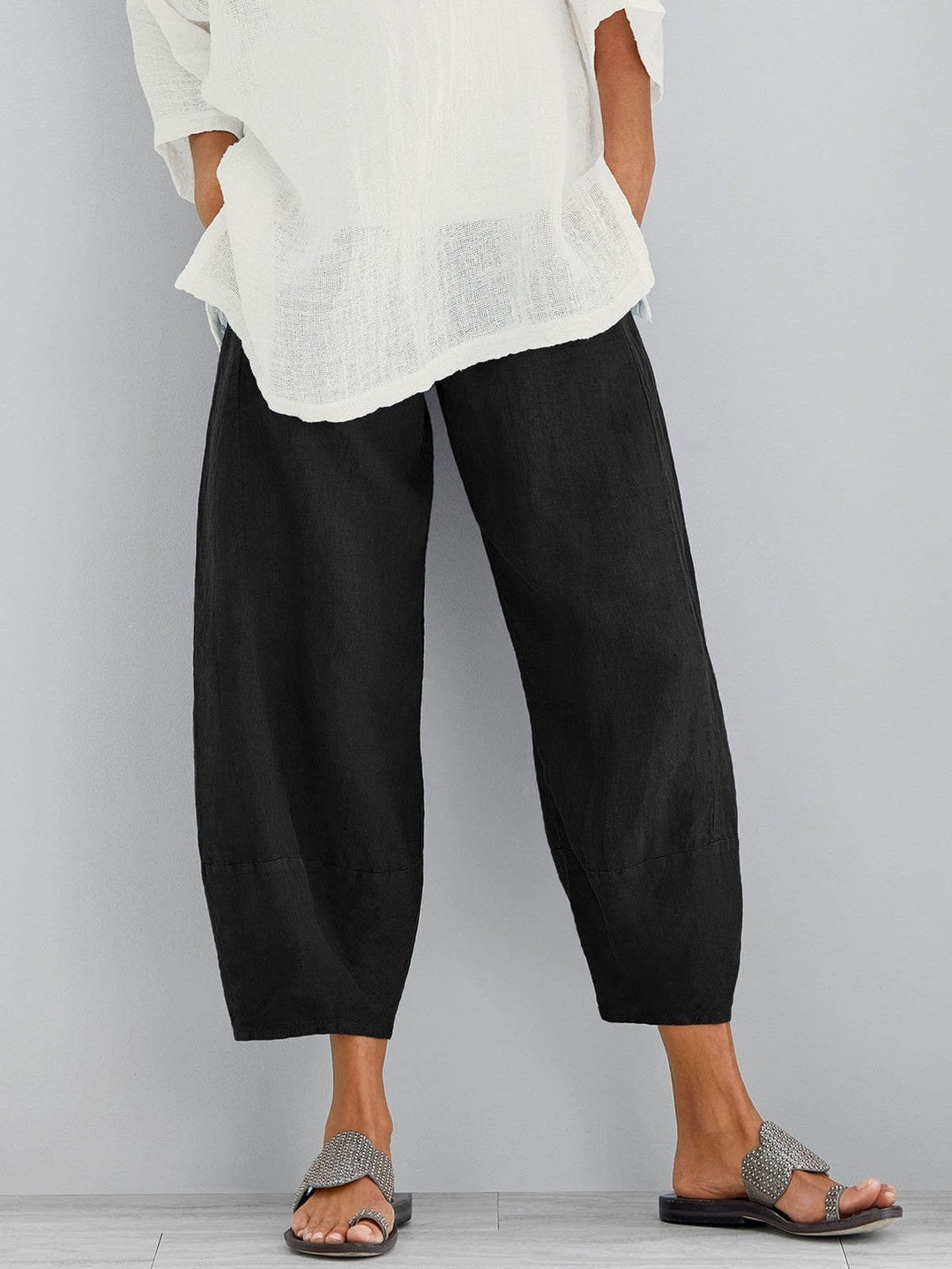 Pantalon large noir à taille élastique et coupe courte, porté avec une blouse blanche aérienne et des sandales sur fond gris. 3