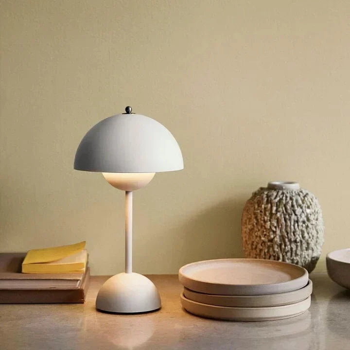 Lampe champignon blanc cassé allumée sur un plan de travail, près de livres, d'assiettes en grès empilées et d'un vase texturé. 1