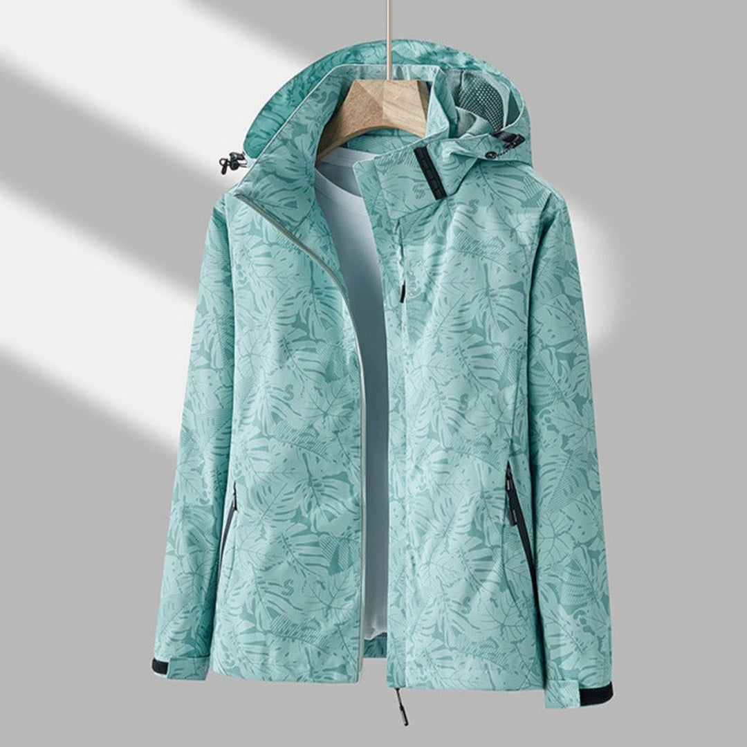 Veste vert menthe à imprimé de feuilles, capuche avec filet d'aération, zip ouvert sur un t-shirt clair, suspendue à un cintre. 7